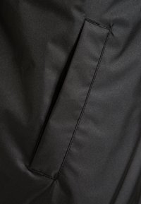 Černá tkanina s hladkým povrchem obsahuje jednu kapsu přišitou na boku, s čistým stehem podél okrajů.