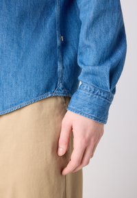 Camicia in denim blu con colletto e polsini con bottoni. Cuciture visibili e un'etichetta su un lato. Parte del tessuto di un pantalone beige è parzialmente visibile.
