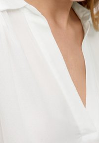 Blusa bianca realizzata in tessuto leggero e leggermente trasparente, con scollatura a V e colletto ripiegato. Texture liscia con un lieve lucido.