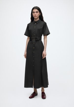 Woman wearing a black short-sleeve buttoned midi dress with a wide belt, paired with burgundy loafers, standing against a plain white background.