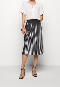 Femme portant un chemisier blanc à manches courtes, une jupe plissée noir et blanc, des sandales à talons hauts noires, tenant un sac à main métallisé.