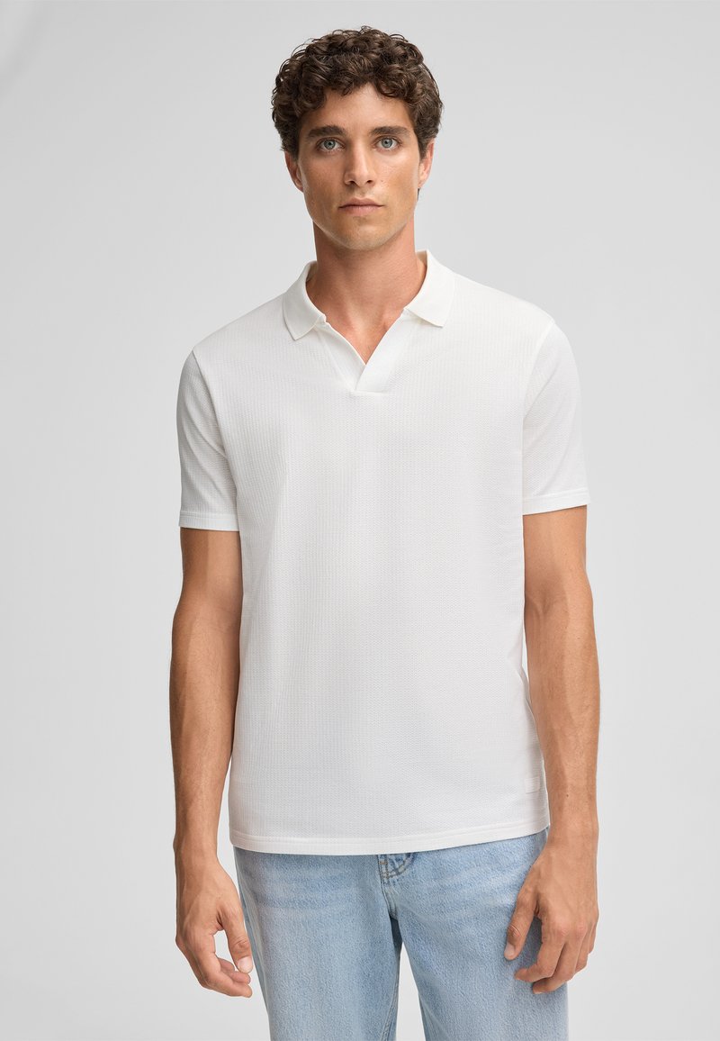 Weißes Poloshirt mit kurzen Ärmeln aus strukturierter Stoff, schmalem Kragen und lockerer Passform. Kombiniert mit hellblauen Jeans.