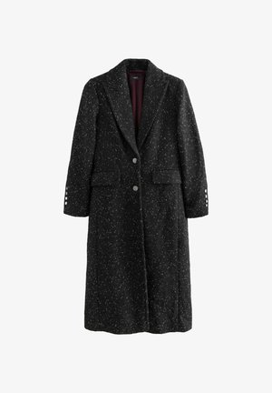 Mujer vistiendo un mini vestido plateado brillante bajo un abrigo negro largo con textura, de pie sobre escalones de mármol dentro de un edificio de piedra.