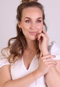Gouden sieraden omvatten een hartvormige hanger ketting, bijpassende oorbellen en een armband met roze accenten, gedragen met een witte top.