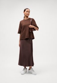 Žena stojí v hnedom huňatom svetri, lesklých hnedých dlhých sukňach, bielych teniskách, s veľkými hooped náušnicami a drží hnedú tašku cez rameno.