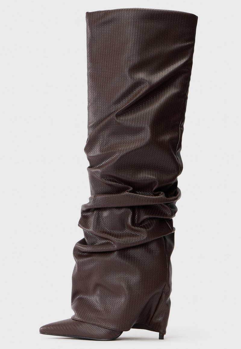 Bota de caña alta marrón oscuro, suelta, con un patrón texturizado y punta afilada sobre un tacón bajo, contra un fondo liso.
