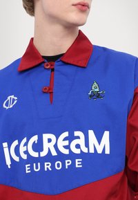 Pullover blu e rosso con colletto bottonato, caratterizzato dal testo "ICECREAM EUROPE" e da un personaggio dei cartoni animati ricamato sul petto.