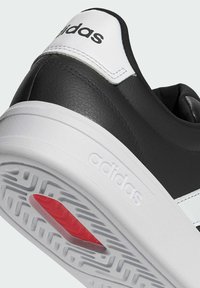 Talonde de sneaker Adidas noir et blanc avec logo embossé sur semelle blanche et détail de grip rouge sur le motif de la semelle.