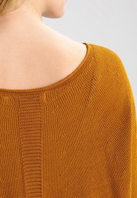 Sinapinkeltainen neulepaita, jossa leveä, pyöreä pääntie. Sisältää tekstuurisen kuvion ja sileän kankaan olkapään ja selän alueella.