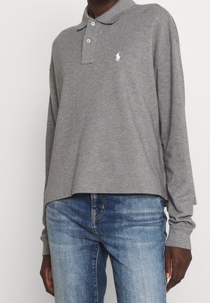 Mężczyzna ubrany w szarą koszulkę polo z długim rękawem z białym logo i niebieskie dżinsy, stojący na jednolitym tle.