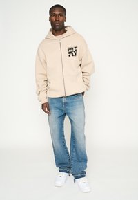 Haut à capuche zippé beige avec un motif noir sur la poitrine, associé à un jean droit bleu clair et des sneakers blanches.