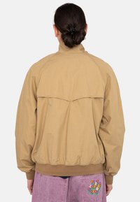 Tan lightweight jacket with a high collar and elastic hem; features a back yoke design. Worn with pink pants displaying colorful patterns.