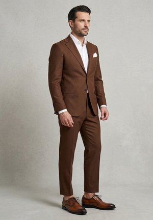 Mand iført brun jakkesæt med hvid skjorte og lommetørklæde, iført brune pæne sko foran ensartet lysegrå baggrund.