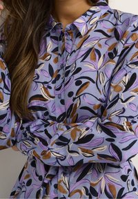 Langarmhemd mit Knopfverschluss in Helllila, das ein Blumenmuster mit schwarzen, braunen und lavendelfarbenen Blättern auf einem weichen Stoff zeigt.