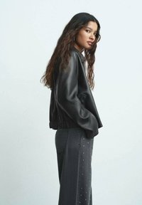Giacca bomber nera in pelle sintetica con vestibilità corta, bordo a costine e tasche laterali, abbinata a jeans scuri a vita alta con dettagli borchiati.