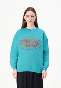 Sweatshirt turquesa com um logo redondo "AMERICAN VINTAGE" em vermelho escuro. Textura suave, corte oversized e punhos canelados. Calças listradas pretas.