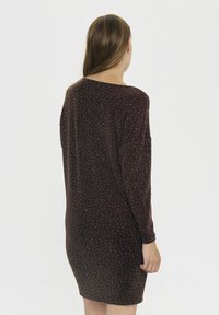 Moteris su ilgais plaukais, vilkinti tamsiai rudą, blizgantį, ilgomis rankovėmis iki kelių drabužį, stovi atsukusi nugarą ant šviesaus fono.