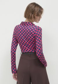 Long-sleeved shirt in navy with large pink polka dots, featuring a collared neckline and buttoned cuffs, paired with brown wide-leg pants.