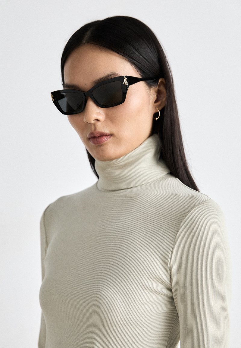 Ochelari de soare dreptunghiulari negri, cu rame groase, evidențiind accente hardware de aur. Modelul poartă un pulover bej deschis, cu guler înalt și textură ribbed.