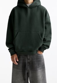 Felpa verde scuro con vestibilità ampia, cappuccio con coulisse, tasca frontale e polsini a coste. Abbinata a jeans larghi grigi sbiaditi.