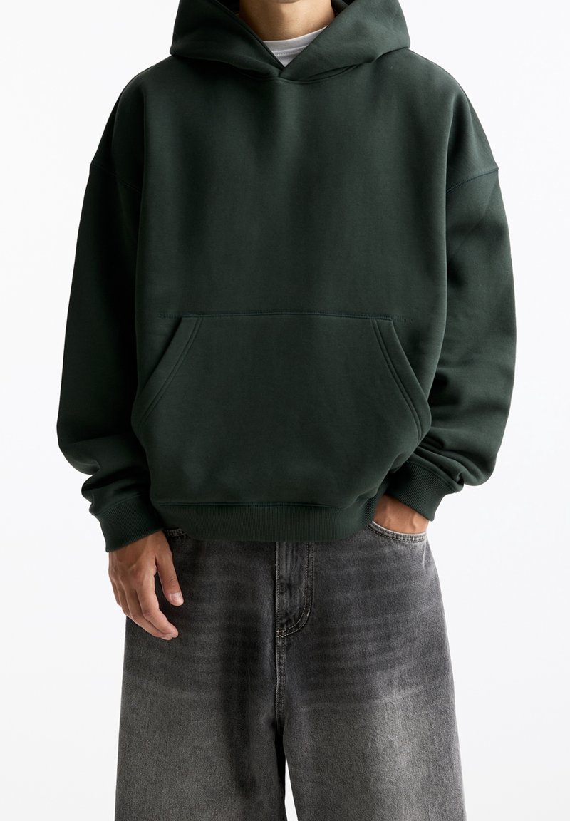 Felpa verde scuro con vestibilità ampia, cappuccio con coulisse, tasca frontale e polsini a coste. Abbinata a jeans larghi grigi sbiaditi.