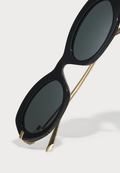 Black oversized sunglasses with dark lenses, featuring a glossy finish and gold metal accents on the temples, with an angular shape.