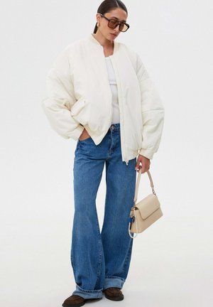 Kobieta w oversize'owej białej kurtce bomberce i szerokich dżinsach, trzyma beżową torbę i okulary przeciwsłoneczne na jednolitym tle.