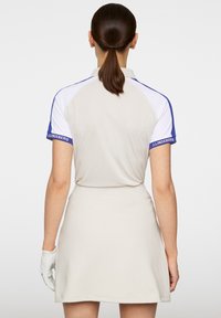 Beige polo-klänning med korta vita ärmar och blå detaljer, med krage och figurnära silhuett. Vit handske delvis synlig.