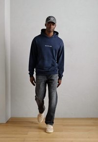 Hanorac bleumarin cu buzunar frontal, logo alb, asortat cu blugi gri și sneakerși de culoare deschisă. Modelul poartă o șapcă gri.