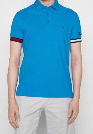 Blauwe polo met een kraag, vijf knoopsluiting en korte mouwen. Voorzien van contrasterende rode en witte strepen aan de mouwomslagen.