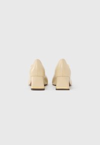 Beige block-heeled shoes with a smooth patent finish, pointed toes, and subtle gold accents on the heel base. Rear view.