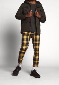 Chaqueta verde oscuro con capucha y múltiples bolsillos, camiseta gris y pantalones de cuadros amarillos y negros. Zapatillas negras y calcetines blancos completan el conjunto.