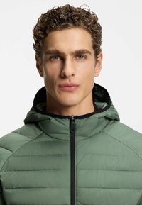 Giovane uomo con capelli ricci, che indossa una giacca di piumino verde con cerniera nera e cappuccio, volto rivolto in avanti su uno sfondo neutro.