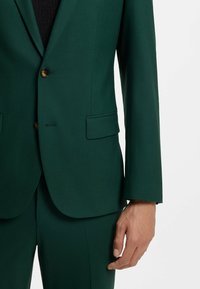 Grüner Blazer aus glattem Stoff, mit einem einzelnen goldenen Knopf, zwei aufgesetzten Taschen und strukturierten Schultern. Taillierte Passform mit klaren Linien.