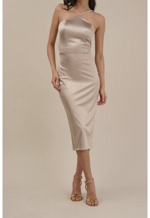 Vrouw draagt een beige satijnen midi-jurk met één glinsterende schouderband en gouden hoge hakken met enkelriempje, staand tegen een effen beige achtergrond.