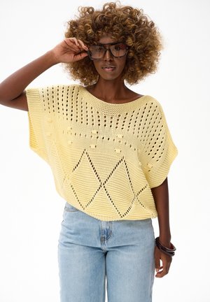 Vrouw met krullend haar die oversized bruine zonnebrillen draagt, een gele gebreide top met ruitpatronen, lichtblauwe spijkerbroek en donkere armbanden.