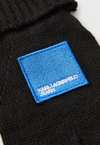 Juodas megztas audinys su siuvinėta mėlyna kvadrato pusele, pažymėta baltais žodžiais „Karl Lagerfeld Jeans“. 