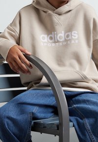 Beige Adidas hoodie med vitt logga, med en känguruficka och ribbade ärmar, kombinerad med blå denimjeans, sittande på en grå bänk.
