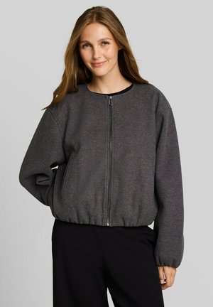 Jeune femme aux longs cheveux portant une veste zippée gris foncé et un pantalon noir, souriant doucement sur un fond gris uni.