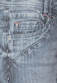 Randiga jeans i denim med blå och vita vertikala linjer. Har en framficka med en silverfärgad metallknapp och röda sömnadspunkter.