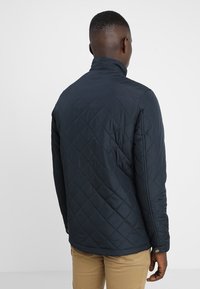 Padded námořnický béžový kabát s vysokým límcem, navržený s kontrastním prošíváním. Vyznačuje se hladkou texturou a zapínáním na knoflíky na manžetách.