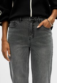 Jeans in denim grigio con vita alta, design classico a cinque tasche e dettagli di cucitura visibili, abbinati a una maglietta nera con accenti bianchi.