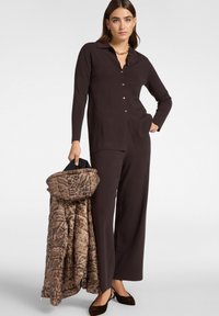 Haut marron à boutons avec manches longues, associé à un pantalon marron ample. Tient un manteau à capuche en imprimé léopard rembourré. Chaussures noires.