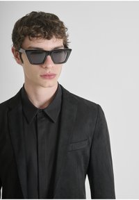 Occhiali da sole neri con una montatura ampia e angolare e lenti oscurate, abbinati a una camicia e a un giubbotto neri, caratterizzati da una leggera trama.