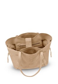 Sac fourre-tout en suède beige avec une large ouverture, poche zippée intérieure et deux longues anses. Présente une texture avec une finition lisse et un branding minimal.