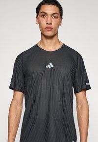 Musta urheilupaita lyhyillä hihoilla, jossa on teksturoitu kuvio, heijastava Adidas-logo ja kapea istuvuus.