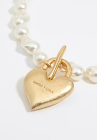 Ciondolo a forma di cuore in oro con incisione "BIMBA Y LOLA", attaccato a una collana caratterizzata da perle bianche di forma irregolare.