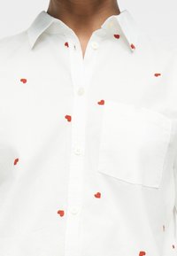 Λευκό πουκάμισο με κουμπιά, διακοσμημένο με κόκκινα κεντημένα καρδιές διάσπαρτες στο μπροστινό μέρος και τσέπη στο στήθος στην αριστερή πλευρά.