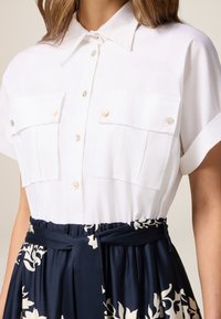 Cămașă albă cu mânecă scurtă, având două buzunare pe piept, cu nasturi la frunte și nasturi multicolori; asortată cu o fustă marine cu un model floral.