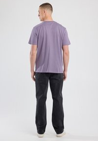 Mann, der mit dem Rücken zur Kamera steht, trägt ein einfaches lila T-Shirt, schwarze Jeans und weiße Turnschuhe vor einem weißen Hintergrund.